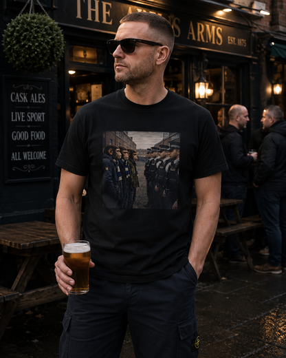 A man wearing a black graphic t-shirt and dark cargo pants stands outside a pub holding a pint of beer. The t-shirt features a rectangular graphic print of soldiers in formation.