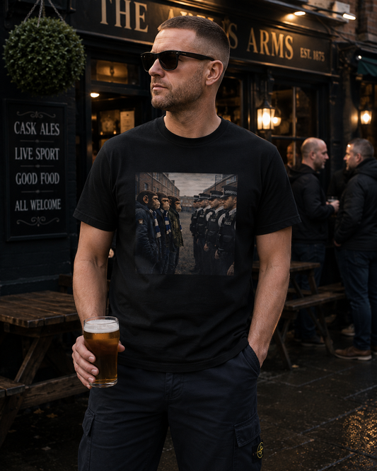 A man wearing a black graphic t-shirt and dark cargo pants stands outside a pub holding a pint of beer. The t-shirt features a rectangular graphic print of soldiers in formation.