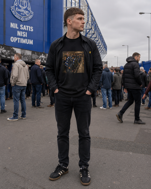 A man wearing a black crewneck sweatshirt with a graphic print, a black zip-up jacket, black jeans, and black sneakers with white stripes, standing in front of an Everton stadium sign.