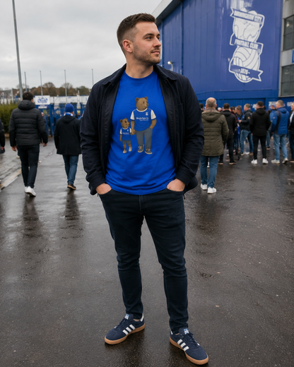 A man wearing a Birmingham t-shirt  with a teddy bear design, a black zip-up jacket, dark jeans, and blue sneakers, standing outdoors in front of a blue building with people in the background.