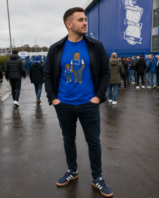 A man wearing a Birmingham t-shirt  with a teddy bear design, a black zip-up jacket, dark jeans, and blue sneakers, standing outdoors in front of a blue building with people in the background.