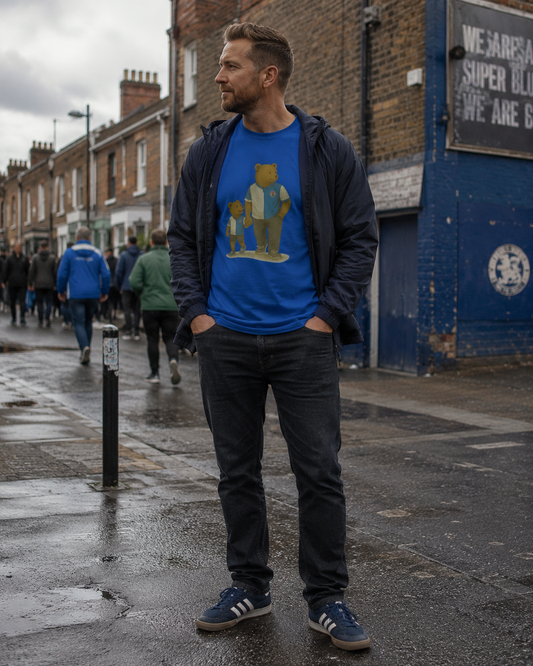A man standing on a wet city street wearing a bright blue graphic T-shirt under a dark navy zip-up jacket, dark jeans, and blue sneakers. The T-shirt features a teddy bear illustration.