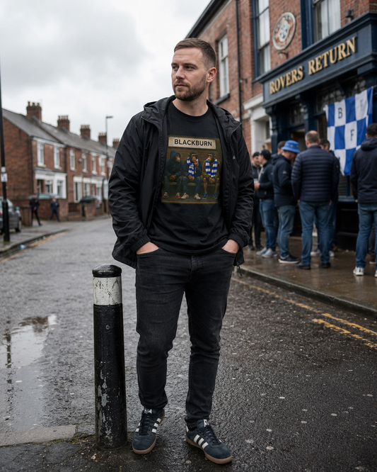 A man standing on a wet street in front of a pub, wearing a black graphic t-shirt with a 'Blackburn' band print, a black hoodie, and dark jeans.