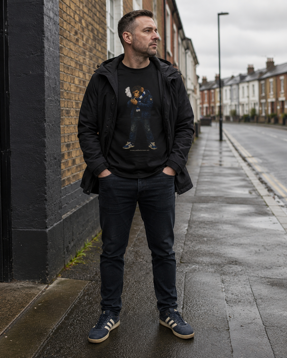 A man stands on a wet city sidewalk wearing a black graphic t-shirt under a black zip-up hoodie, dark jeans, and navy blue sneakers with white stripes.