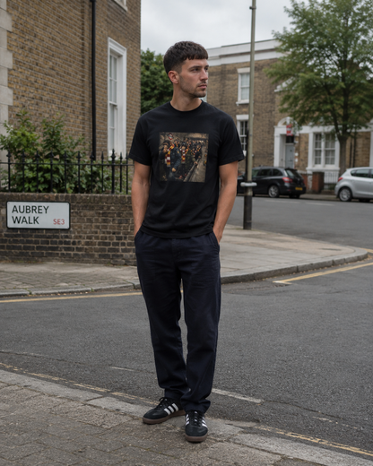 A man standing on a street corner wearing a black graphic t-shirt, dark navy trousers, and black sneakers with white stripes. The background shows a residential street with brick houses, a street sign reading 'AUBREY WALK', and parked cars.