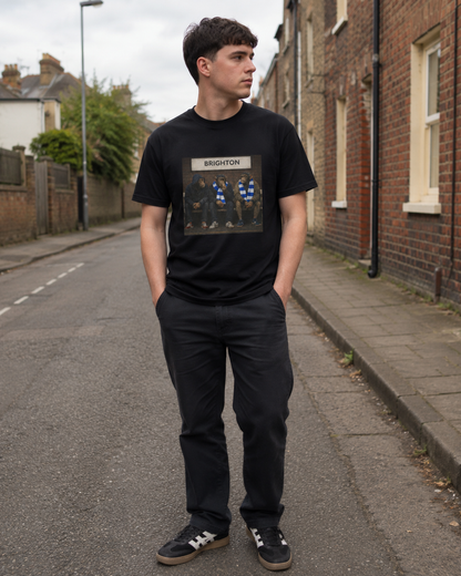A man standing on a street wearing a black Brighton t-shirt with a Brighton graphic on the front, paired with dark trousers and sneakers.