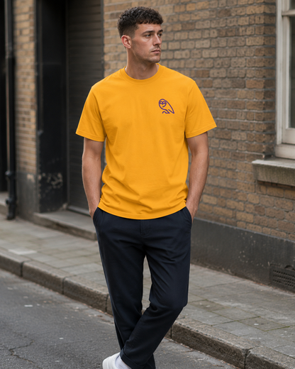 A man wearing a bright yellow SWFC t-shirt with a small embroidered owl logo on the left chest, paired with dark trousers and white sneakers, standing on a city street.