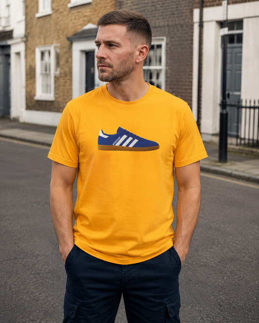 A man wearing a bright yellow SWFC t-shirt with a blue and white sneaker graphic on the chest, standing on an outdoor street with brick houses in the background.