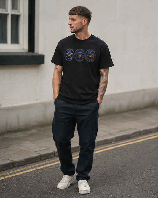 A man wearing a black crew-neck SWFC t-shirt with three circular blue and gold graphic designs across the chest, paired with dark trousers and white sneakers, standing on a street against a light-colored wall.