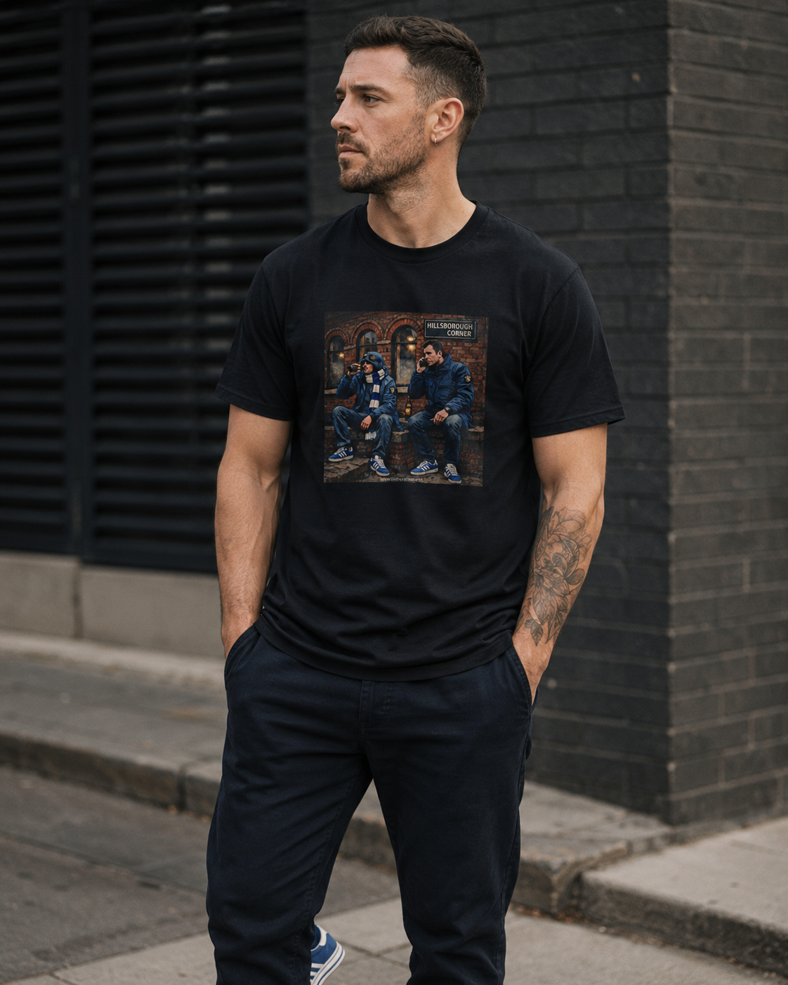 A man wearing a black crewneck SWFC t-shirt with a square graphic print of a street scene, standing outdoors against a dark brick wall.