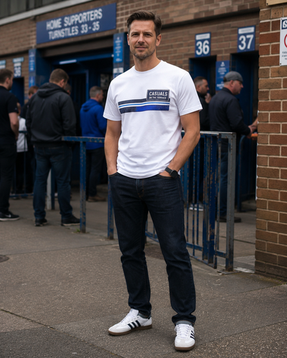A man wearing a white short-sleeve t-shirt with a blue and black Casuals logo across the chest, paired with dark jeans and white sneakers, standing outside a brick building entrance.