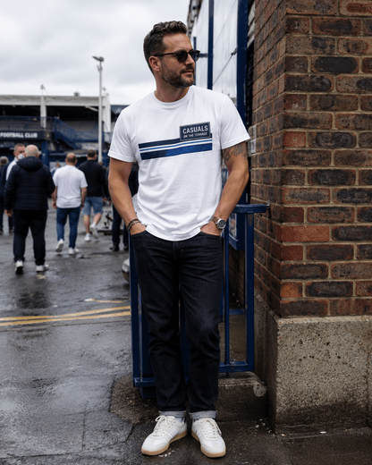 A man wearing a white short-sleeve t-shirt with a blue horizontal stripe and 'CASUALS' logo, paired with dark jeans and white sneakers, standing outdoors next to a brick wall.