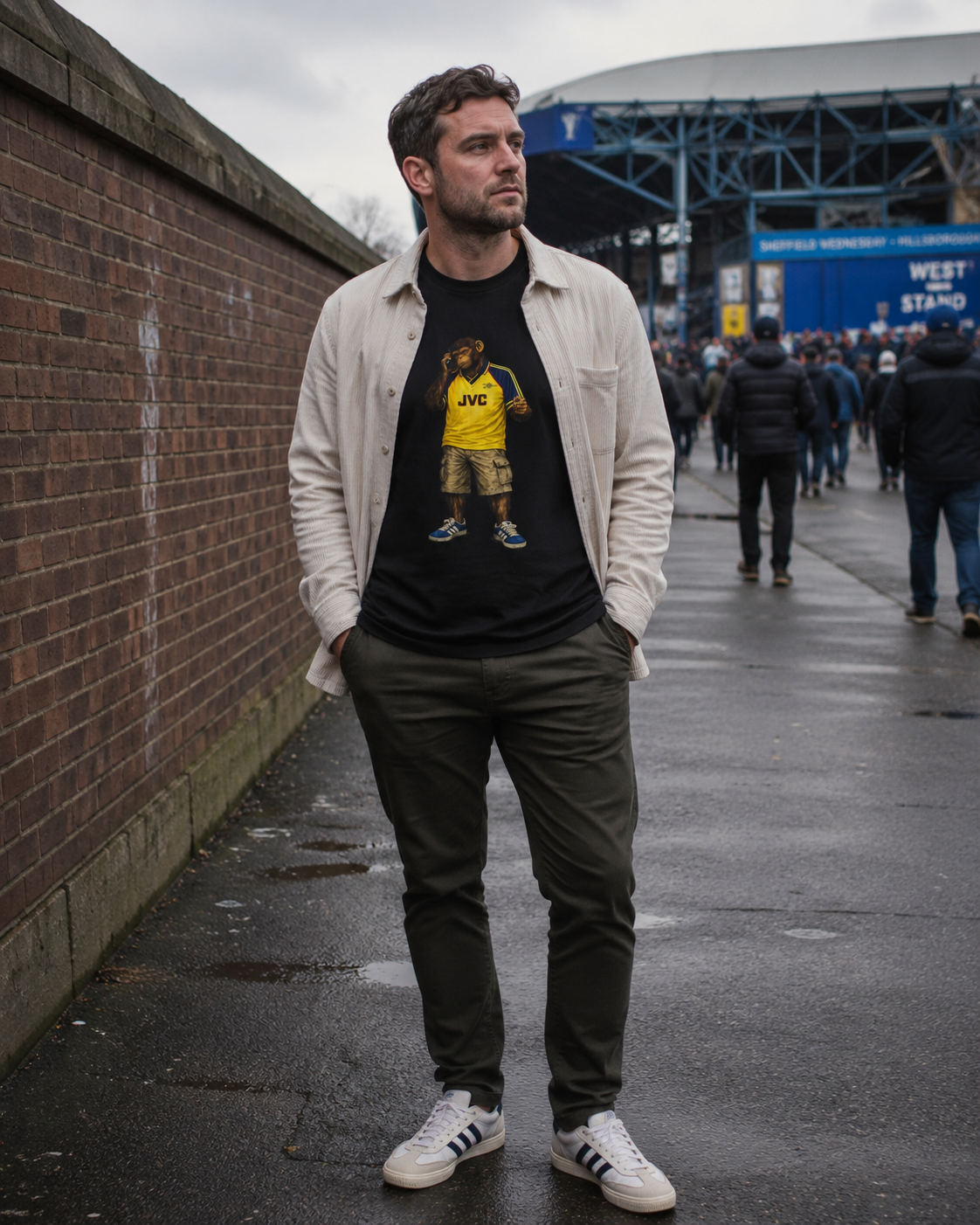 A man standing outdoors on a wet street in front of a brick wall and stadium, wearing a black Arsenal graphic t-shirt under an off-white button-up shirt, dark trousers, and white sneakers.