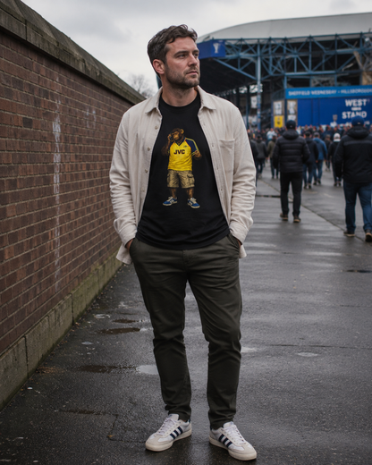 A man standing outdoors on a wet street in front of a brick wall and stadium, wearing a black Arsenal graphic t-shirt under an off-white button-up shirt, dark trousers, and white sneakers.