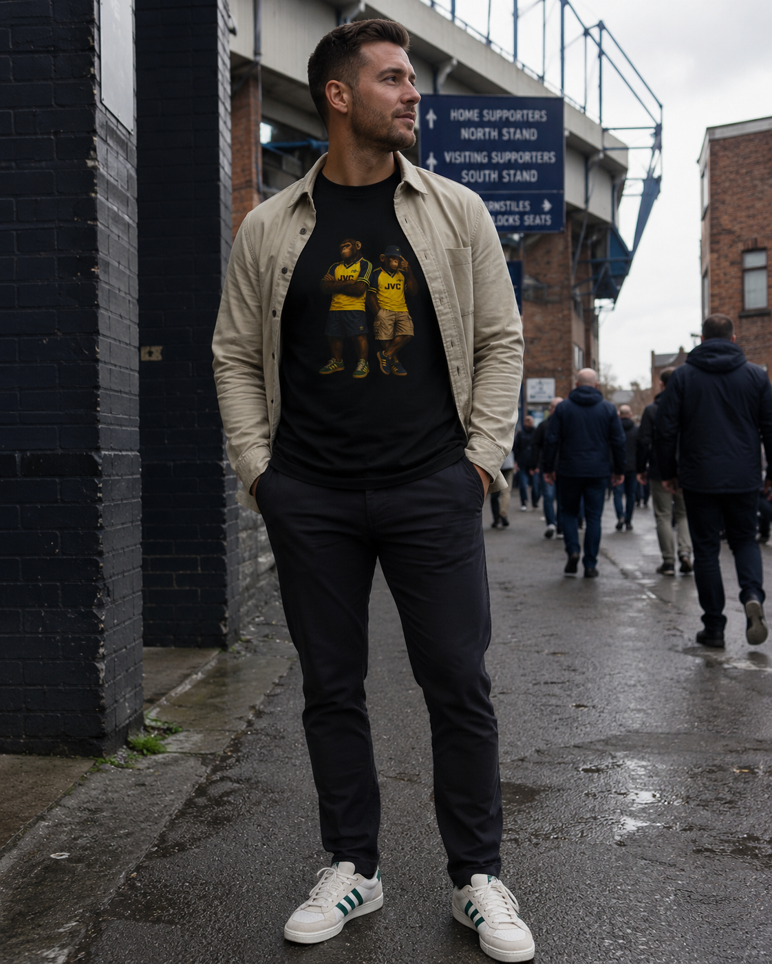 A man wearing a black Arsenal graphic t-shirt under an open beige overshirt, paired with dark trousers and white sneakers, standing on a wet city street.