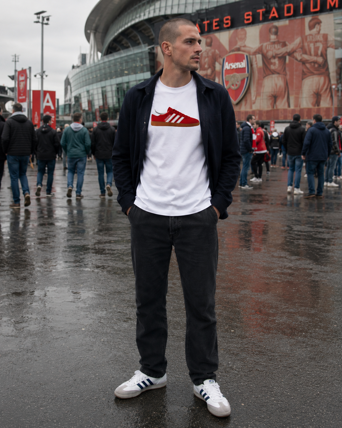 A man wearing a white t-shirt with a red Arsenal Adidas logo, a black zip-up jacket, dark jeans, and white sneakers, standing in front of Arsenal Stadium on a wet, overcast day.
