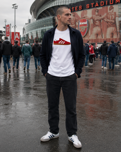 A man wearing a white t-shirt with a red Arsenal Adidas logo, a black zip-up jacket, dark jeans, and white sneakers, standing in front of Arsenal Stadium on a wet, overcast day.