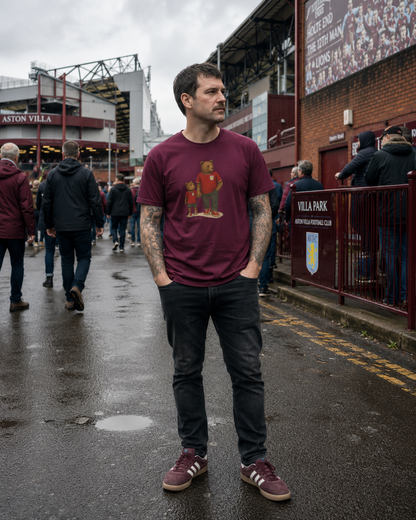 A man standing on a wet street outside Aston Villa Stadium wearing a maroon graphic t-shirt with a red and gold player illustration, paired with dark jeans and matching maroon sneakers.