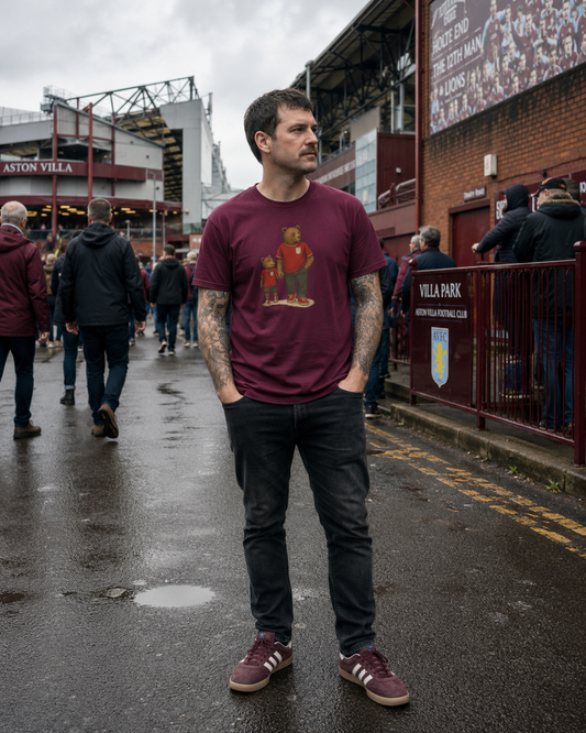A man standing on a wet street outside Aston Villa Stadium wearing a maroon graphic t-shirt with a red and gold player illustration, paired with dark jeans and matching maroon sneakers.