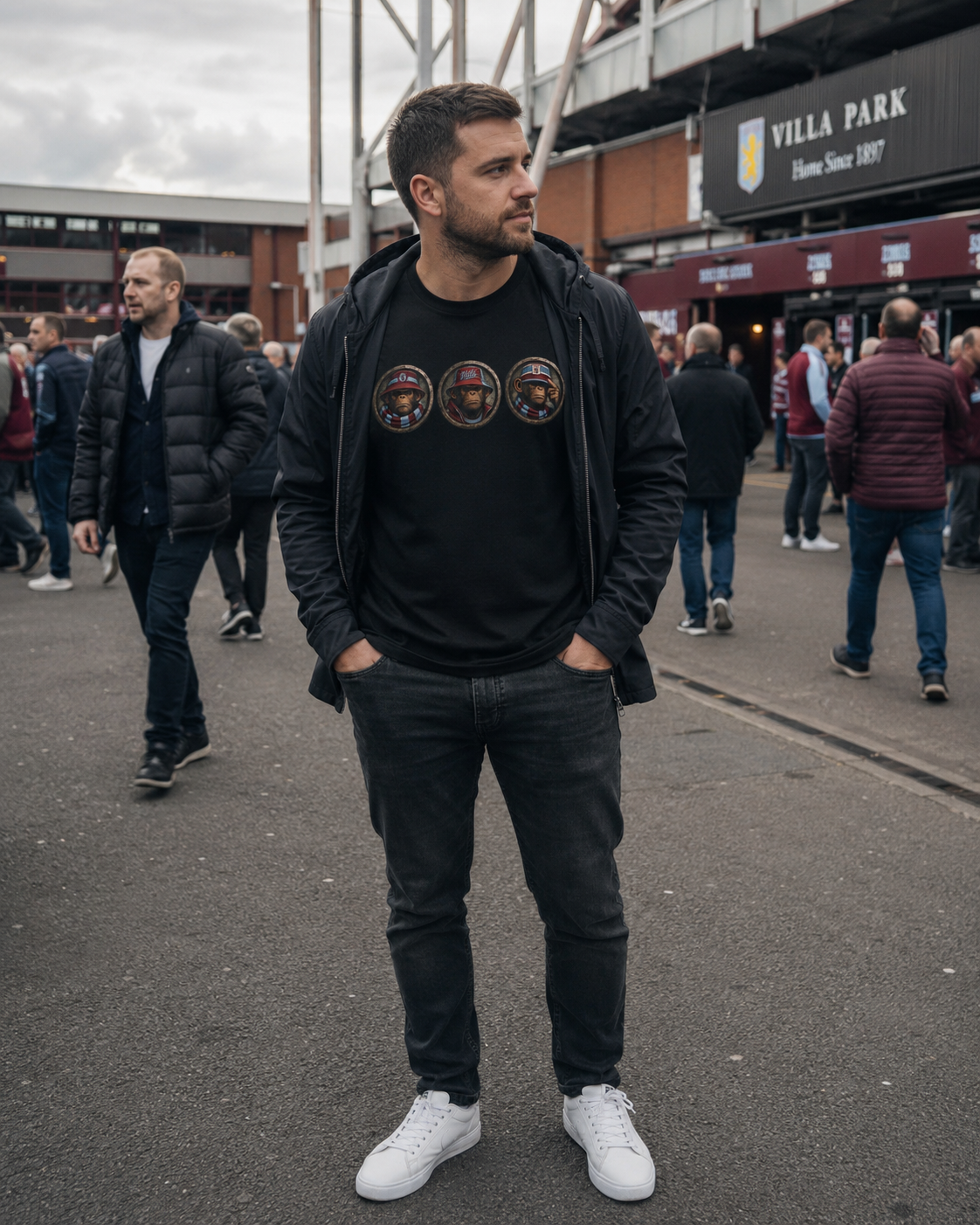 A man wearing a black Aston Villa graphic t-shirt with three circular club crests, a black zip-up hoodie, and black jeans, standing outdoors at Villa Park stadium with other people and the stadium structure in the background.