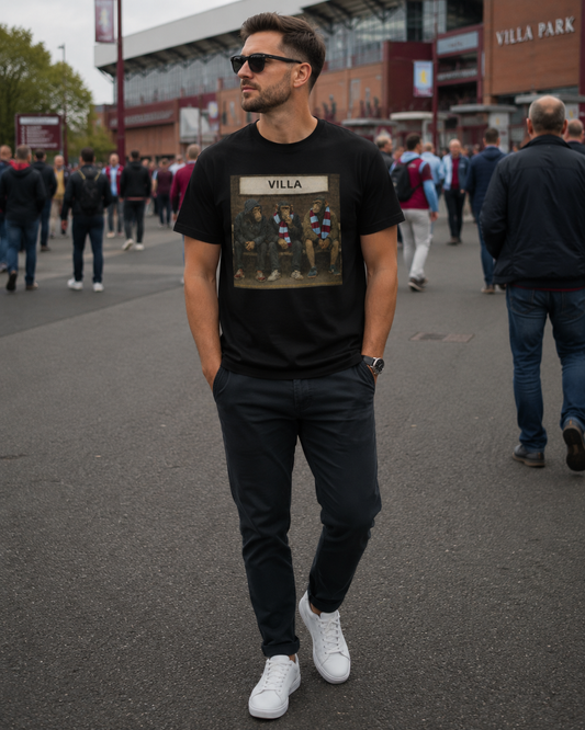 A man wearing a black Aston Villa graphic t-shirt with a vintage-style football team print, dark trousers, and white sneakers, standing in a crowded outdoor street in front of Villa Park stadium.