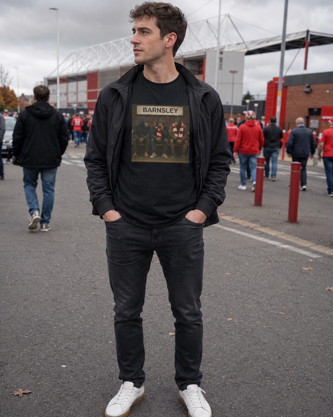 A man wearing a black zip-up jacket over a black t-shirt with a Barnsley graphic, paired with black jeans and white sneakers, standing in front of a stadium in the rain.