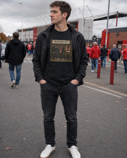 A man wearing a black zip-up jacket over a black t-shirt with a Barnsley graphic, paired with black jeans and white sneakers, standing in front of a stadium in the rain.