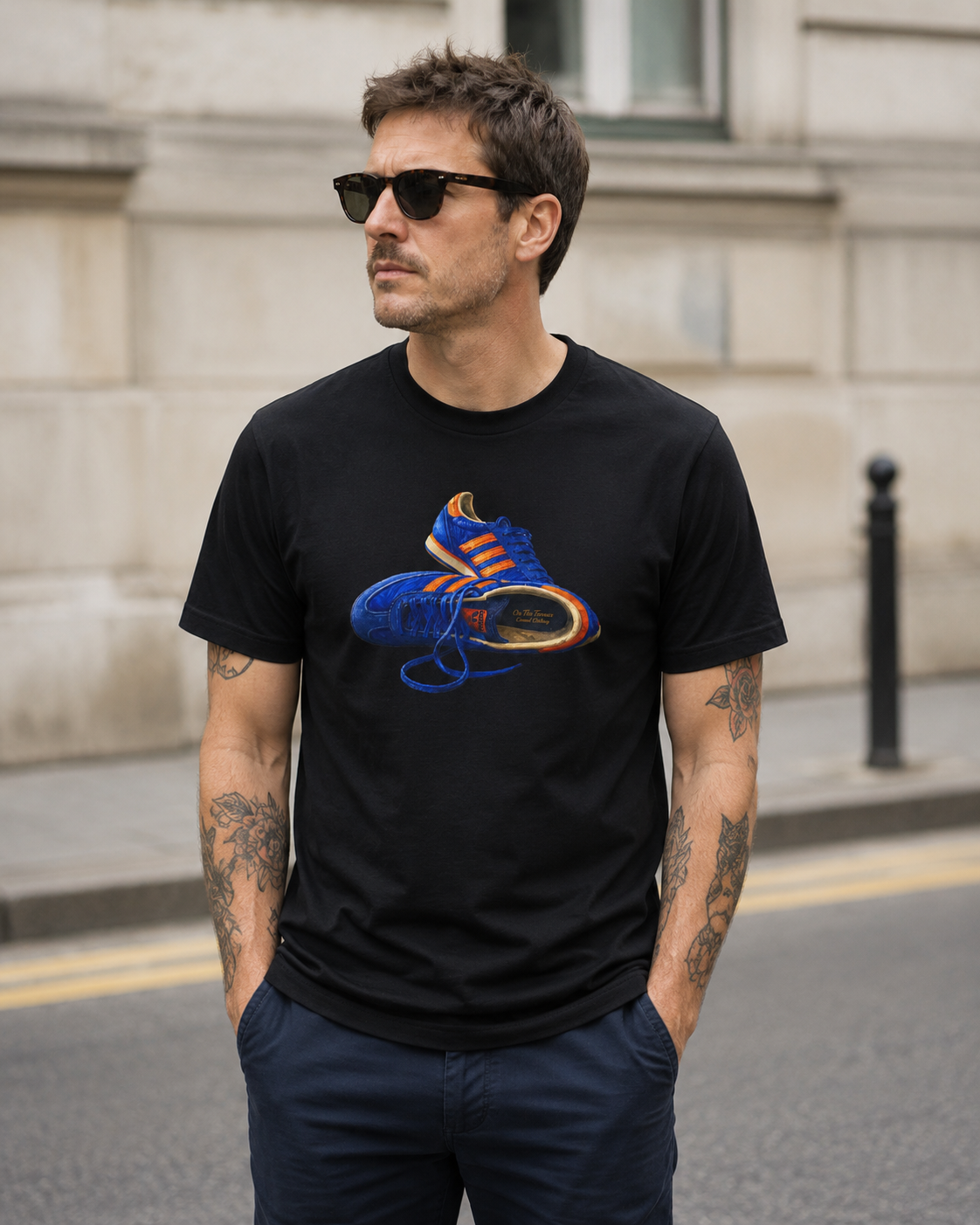 A man wearing a black crewneck t-shirt with a graphic of a blue and orange sneaker, standing outdoors on a city street.