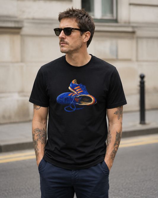 A man wearing a black crewneck t-shirt with a graphic of a blue and orange sneaker, standing outdoors on a city street.