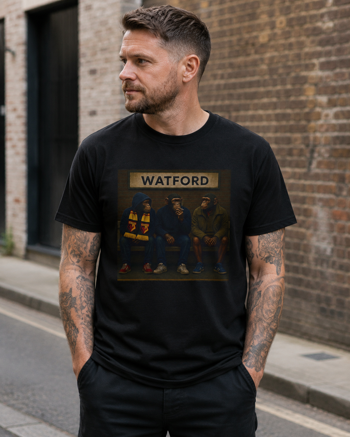 A man wearing a black Watford t-shirt with a Watford-themed print, standing on a city street against a brick wall background.