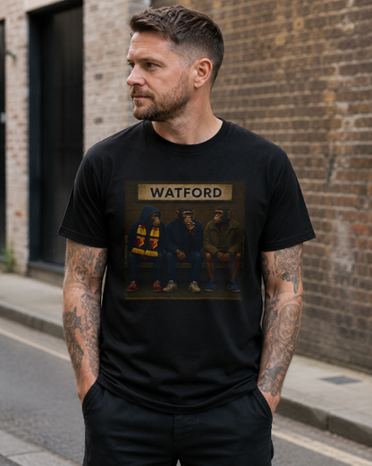 A man wearing a black Watford t-shirt with a Watford-themed print, standing on a city street against a brick wall background.