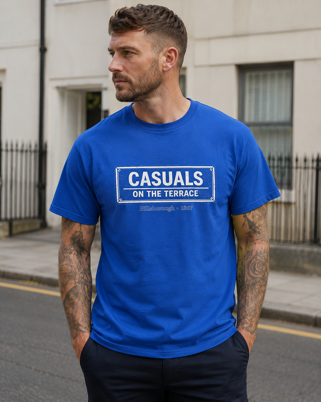 A man wearing a royal blue heavy cotton  SWFC t-shirt flat lay featuring a central vintage blue and white street sign graphic reading "CASUALS ON THE TERRACE" with "Hillsborough • 1867" printed below, and an inner neck label for On The Terrace, standing outdoors on a city street.