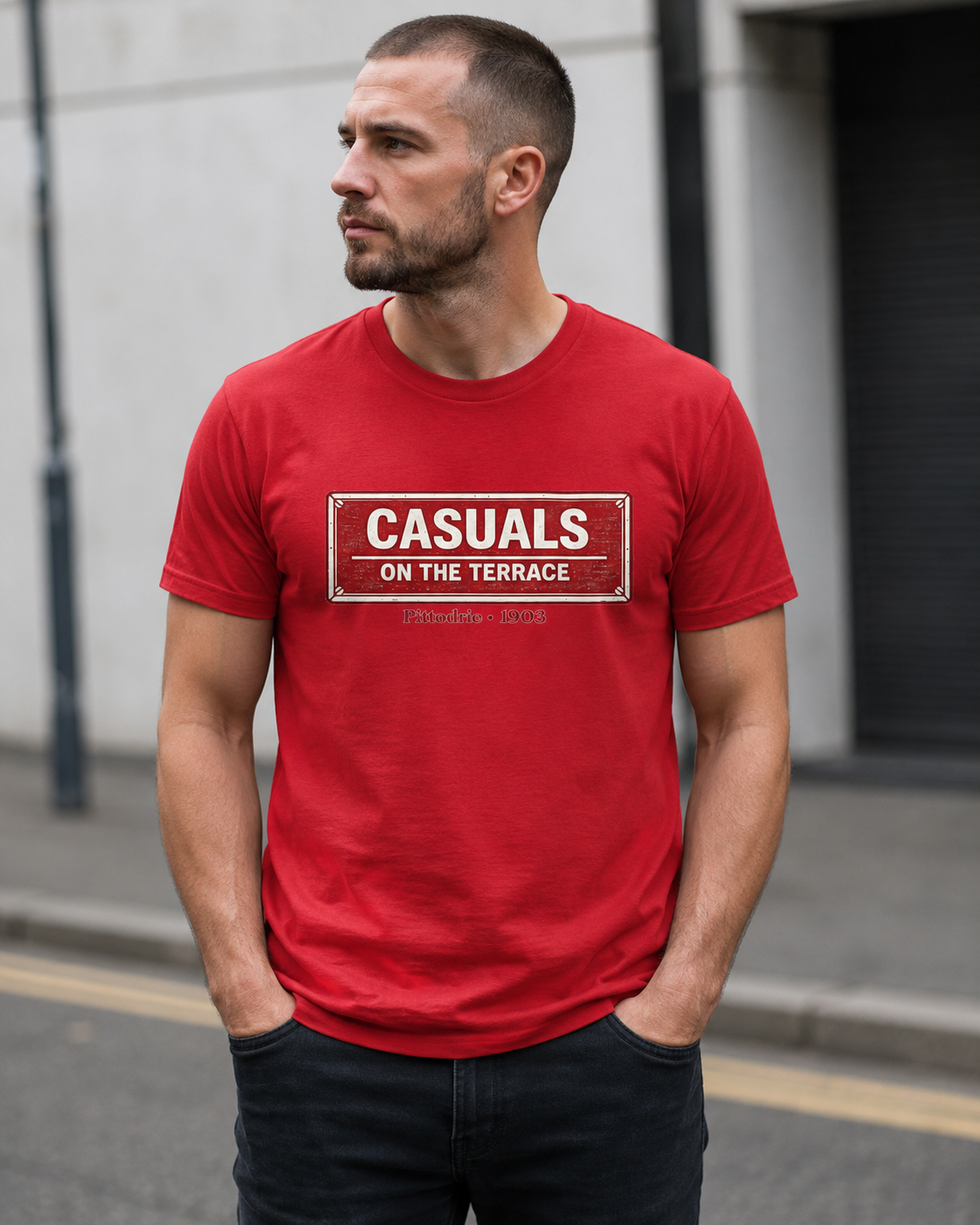 A man wearing a red heavy cotton Aberdeen t-shirt flat lay featuring a central vintage red and white street sign graphic reading "CASUALS ON THE TERRACE" with "Pittodrie • 1903" printed below, and an inner neck label for On The Terrace.', standing outdoors on a city street.