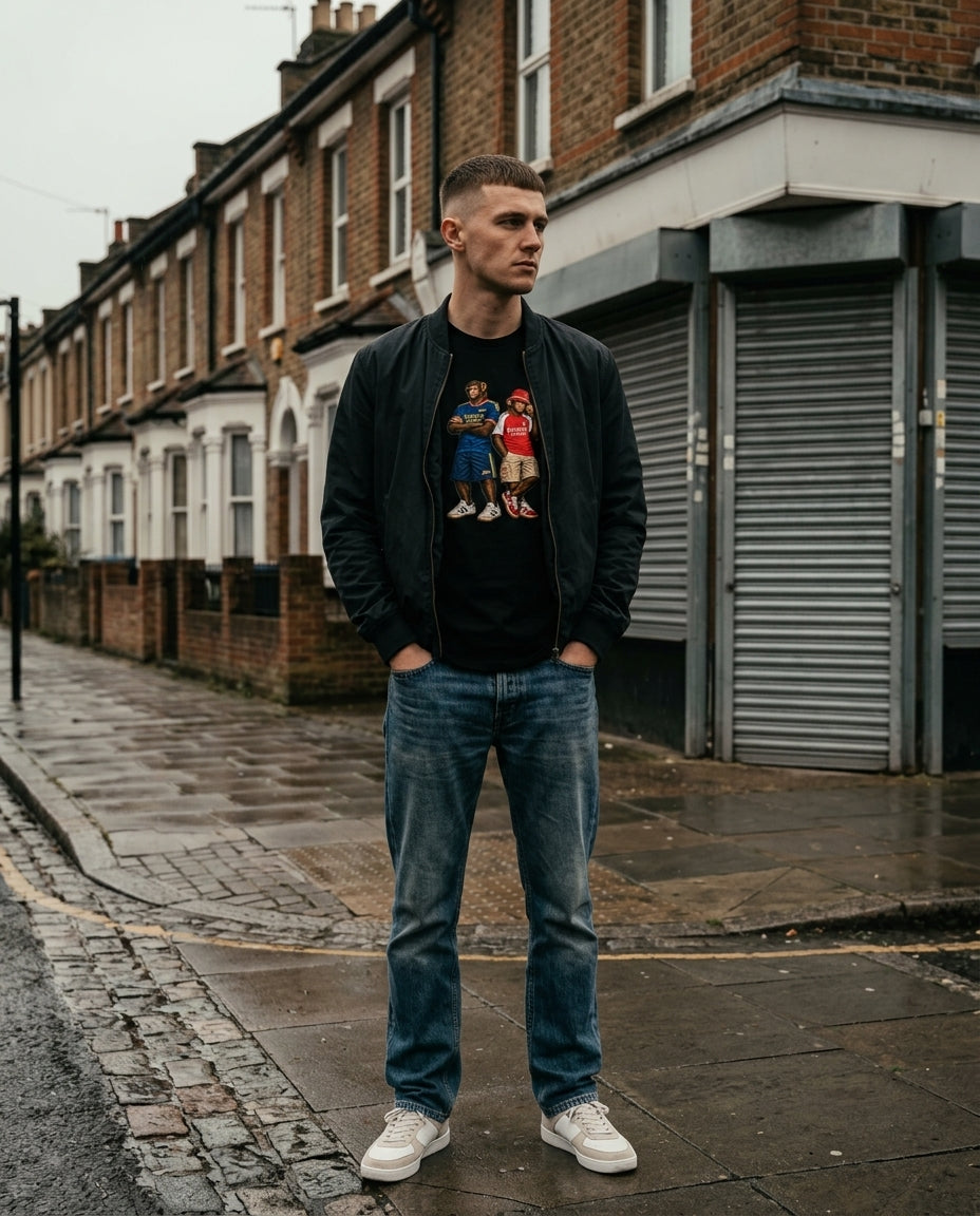 A man stands on a wet cobblestone street wearing a black bomber jacket over a Arsenal Emirates Era Duo t-shirt in black styled with denim - On The Terrace casual wear