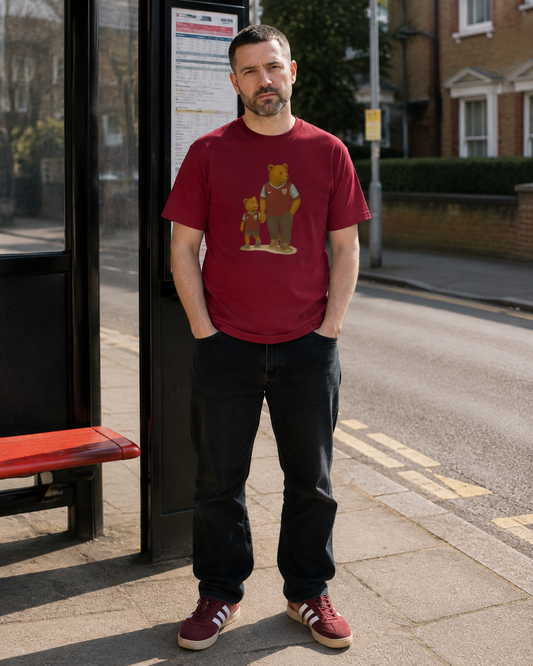 A man standing on a city street next to a bus stop, wearing a maroon crew-neck Burnley t-shirt with a cartoon bear graphic, dark jeans, and maroon sneakers.