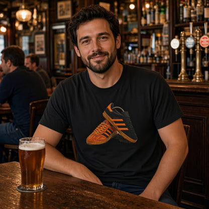 A man sitting at a wooden bar in a pub, wearing a black t-shirt with a graphic of a brown and orange sneaker. A glass of beer is on the table in the foreground, with blurred bar patrons and shelves in the background. Wearing a Terrace fashion tee in black with an orange classic trainer graphic