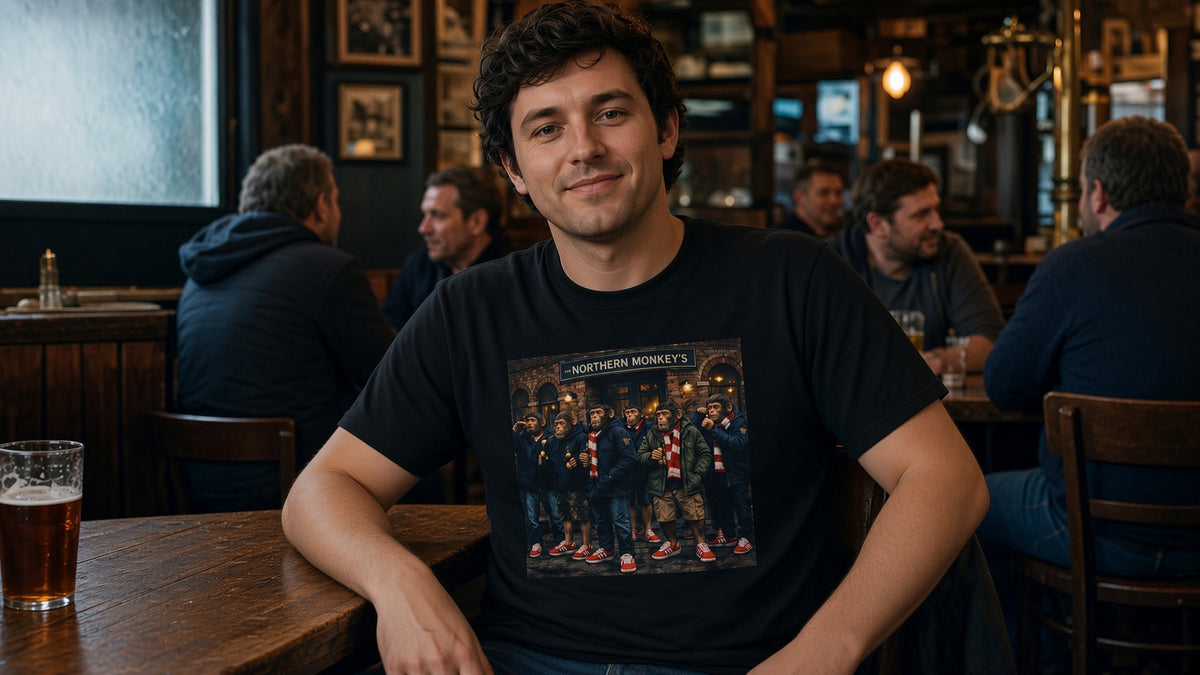 A man sitting at a wooden bar table in a dimly lit pub, wearing the Northern Monkeys 'Red and Whites' tee - chimps outside a brick pub