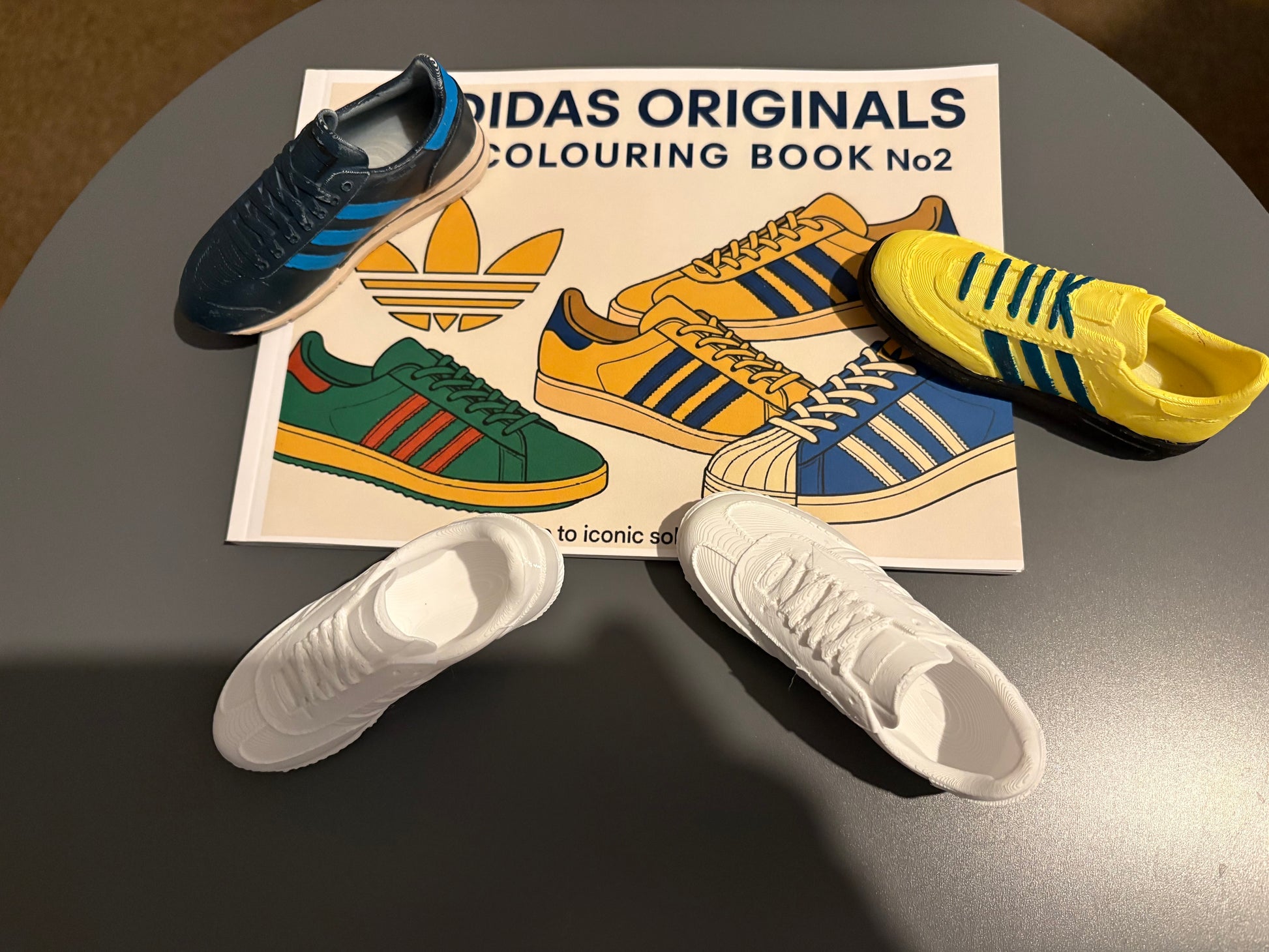 Miniature Adidas sneakers in blue, yellow, and white on a grey table with an Adidas Originals Colouring Book