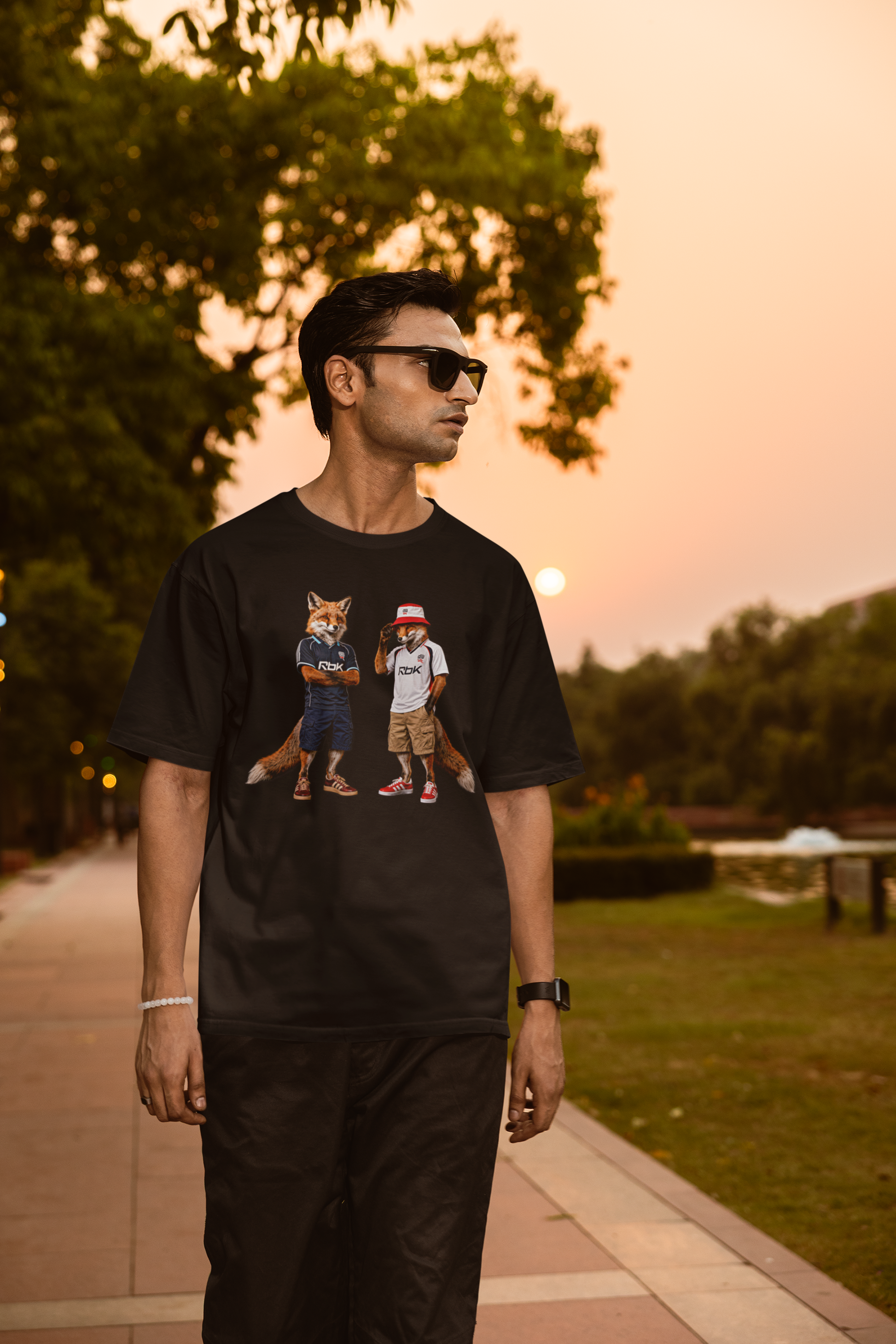 A black graphic t-shirt laid flat, featuring a central design by On The Terrace for Bolton fans. The graphic showcases two anthropomorphic English foxes dressed in vintage Rbk Bolton-inspired football kits. The fox on the left wears the dark blue retro away jersey with arms crossed, while the fox on the right sports the classic white home shirt, a red and white bucket hat, and cargo shorts. The inner neck of the tee features the official "On The Terrace" brand logo