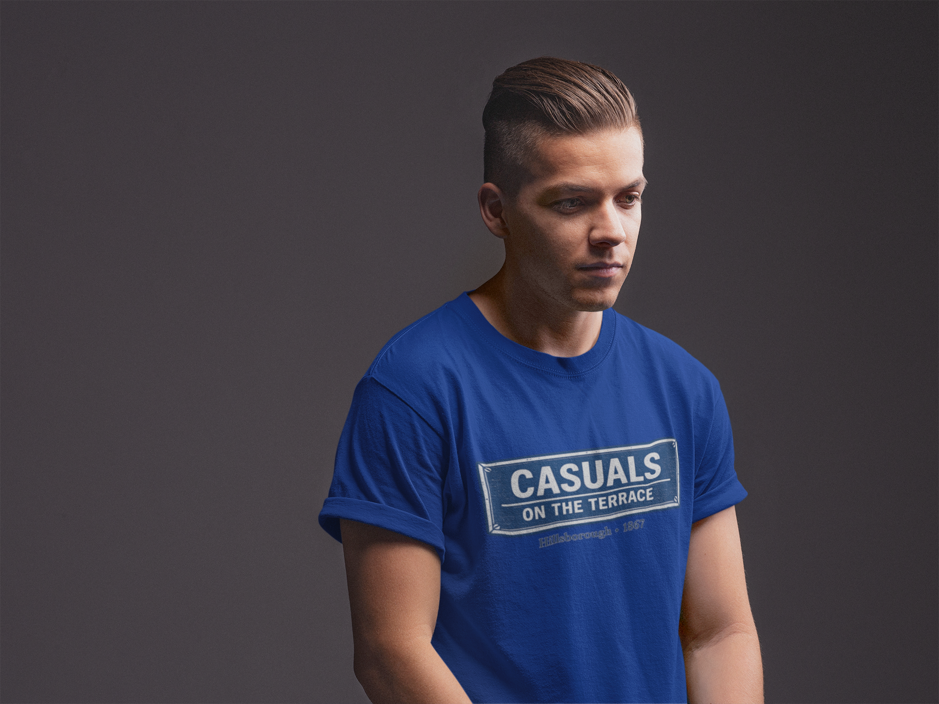 A man wearing a royal blue heavy cotton  SWFC t-shirt flat lay featuring a central vintage blue and white street sign graphic reading "CASUALS ON THE TERRACE" with "Hillsborough • 1867" printed below, and an inner neck label for On The Terrace. The photo is taken against a solid dark gray studio background.