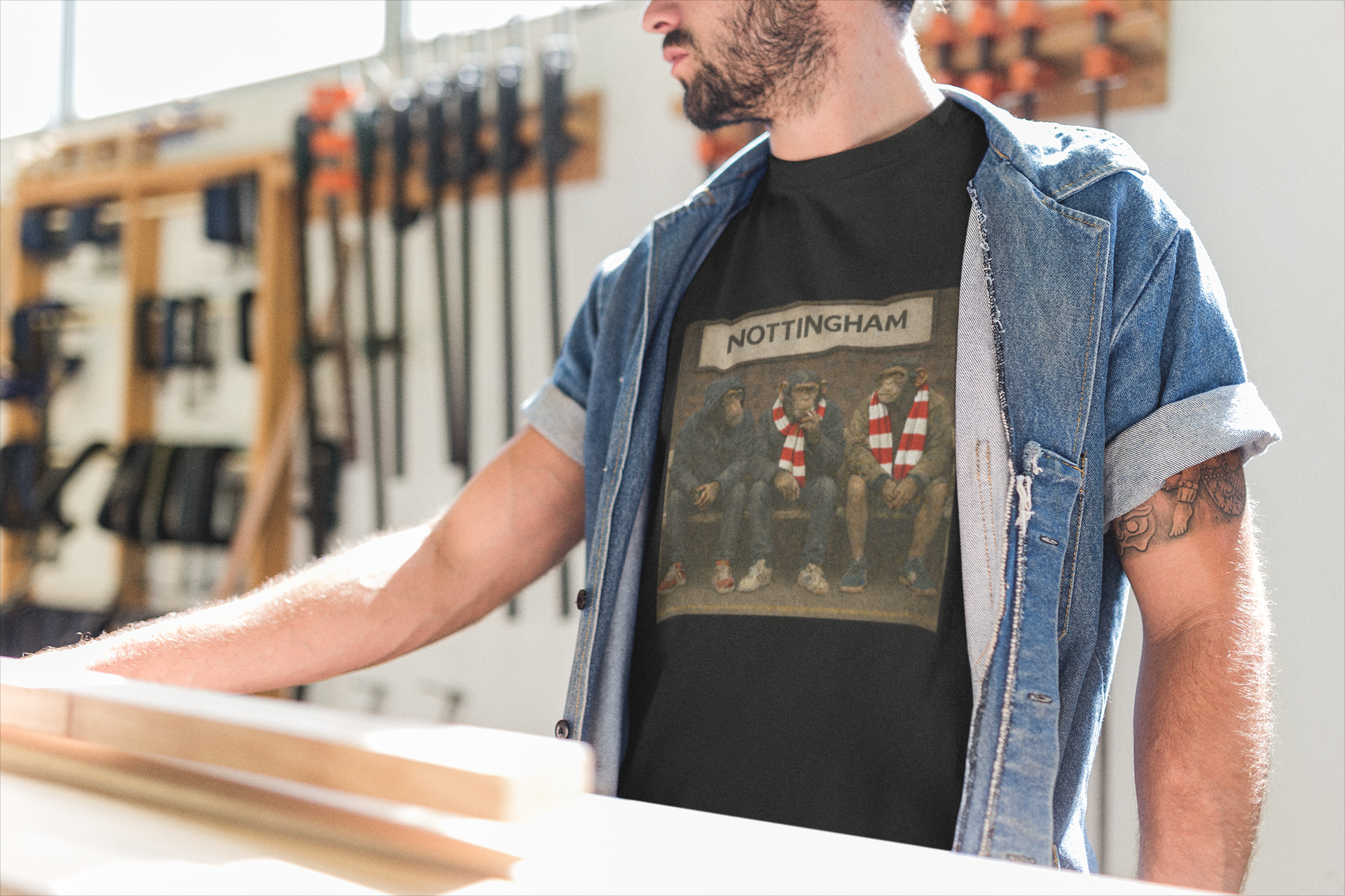 A man wearing a black graphic Notts Forest t-shirt with a Nottingham print, layered under an open blue denim shirt, standing in a workshop environment.