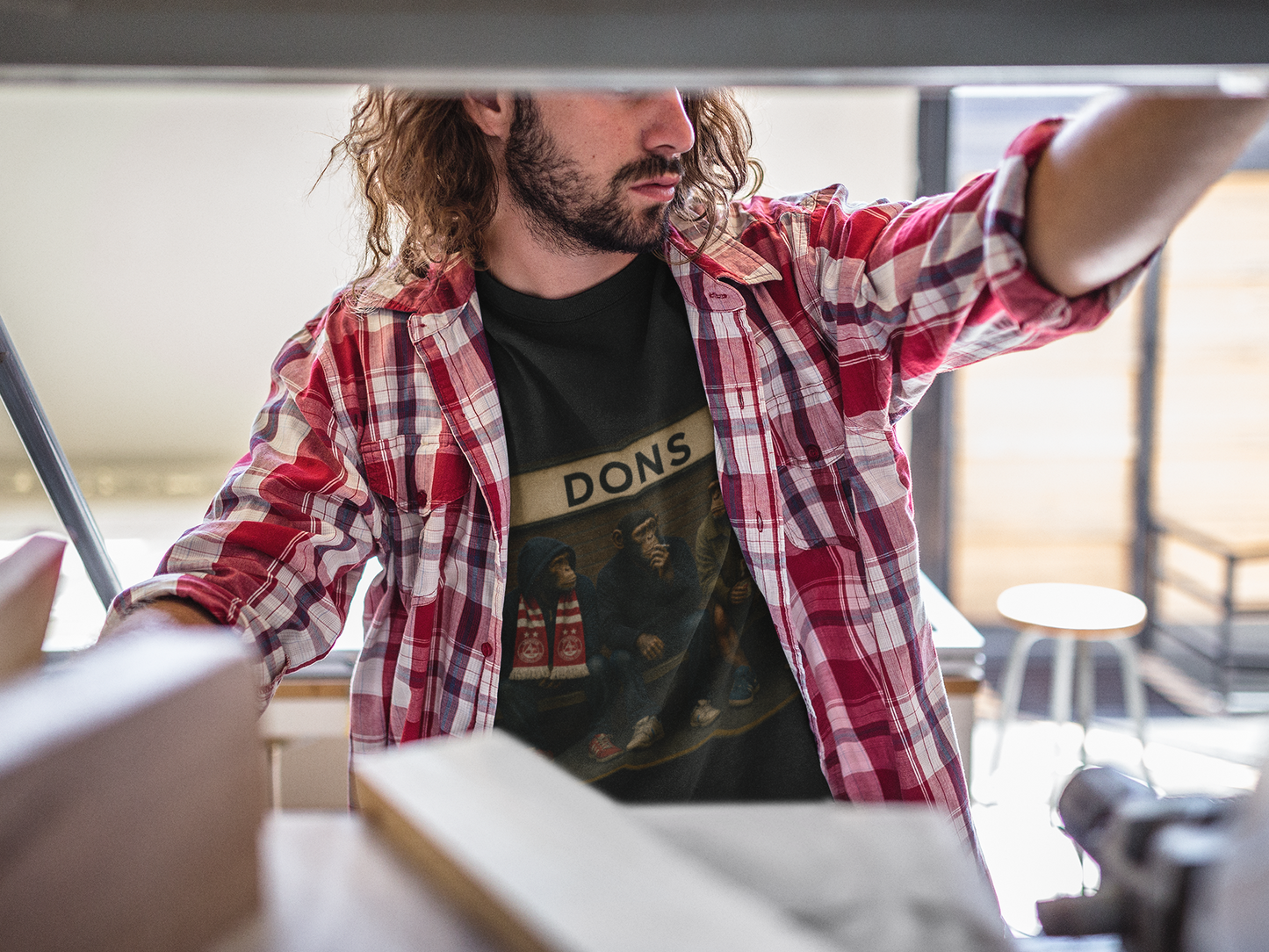 A man with a beard and long hair wearing a red and white plaid flannel shirt over a black graphic Aberdeen t-shirt with 'DONS' printed on the chest, working in a workshop environment.
