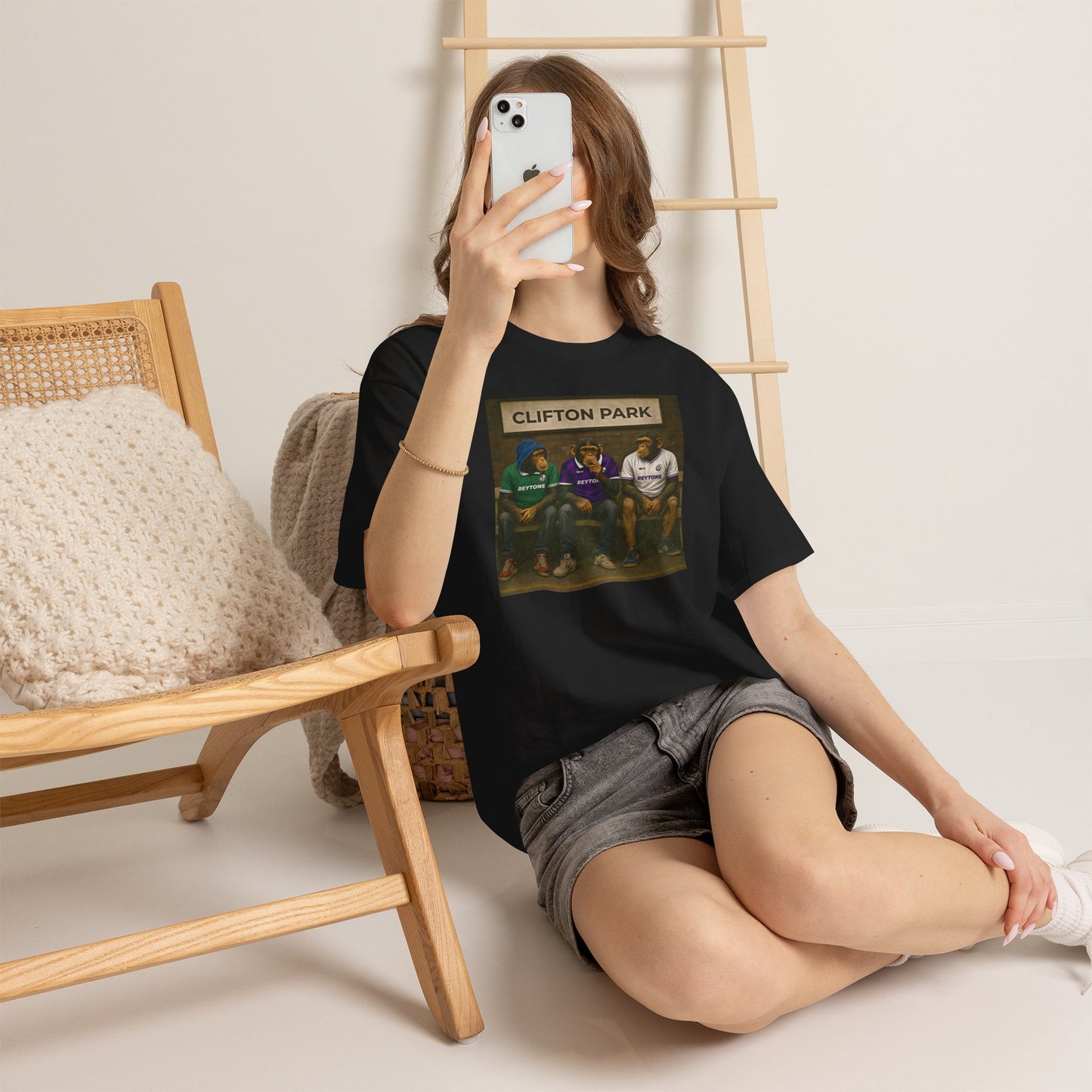 Person wearing a black Reytons t-shirt with a graphic design, sitting on the floor in a room with wooden furniture.