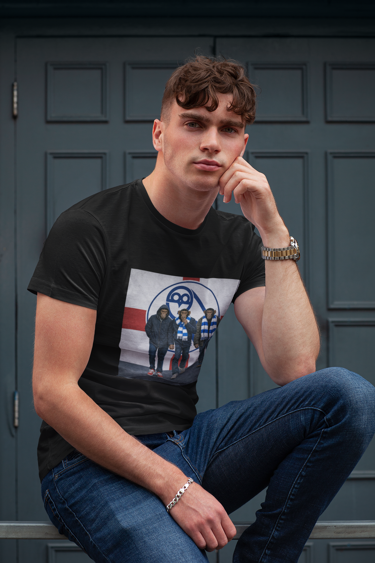 A man wearing a black graphic swfc t-shirt with a band print and blue jeans, sitting in front of a dark paneled door.