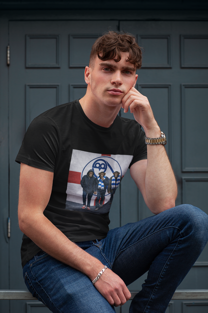A man wearing a black graphic swfc t-shirt with a band print and blue jeans, sitting in front of a dark paneled door.
