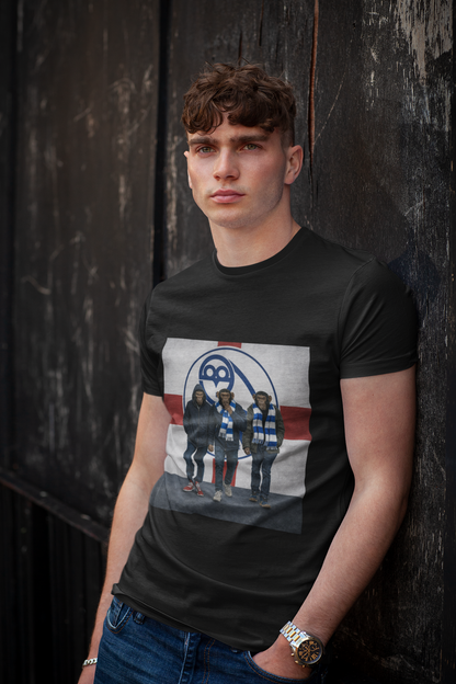 A young man with curly brown hair wearing a black graphic swfc t-shirt and blue jeans, leaning against a dark wooden wall.