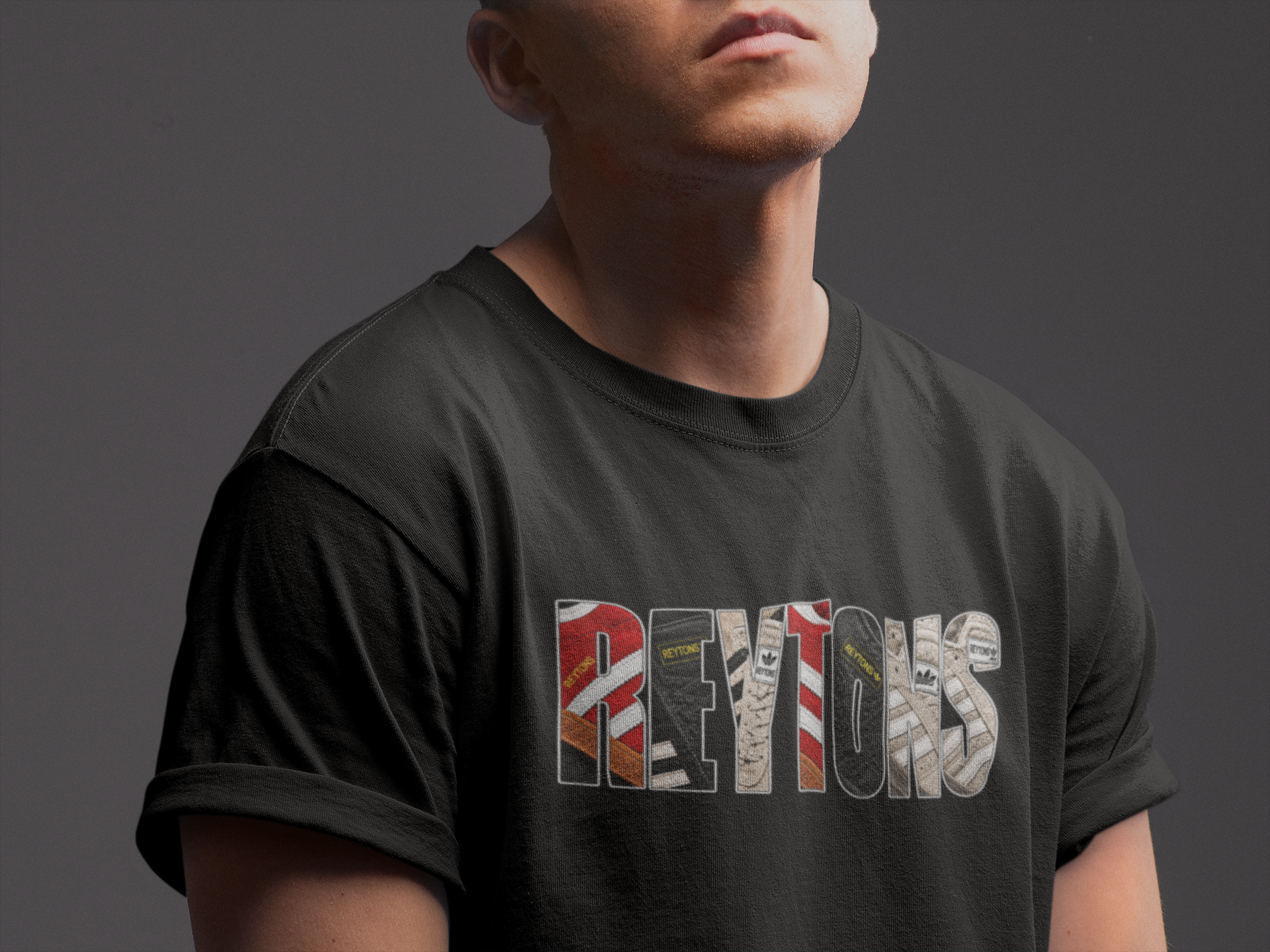 A man wearing a black Reytons t-shirt with a graphic print spelling 'REYTONS' filled with cigarette pack designs, shown from the chest up against a dark gray background.