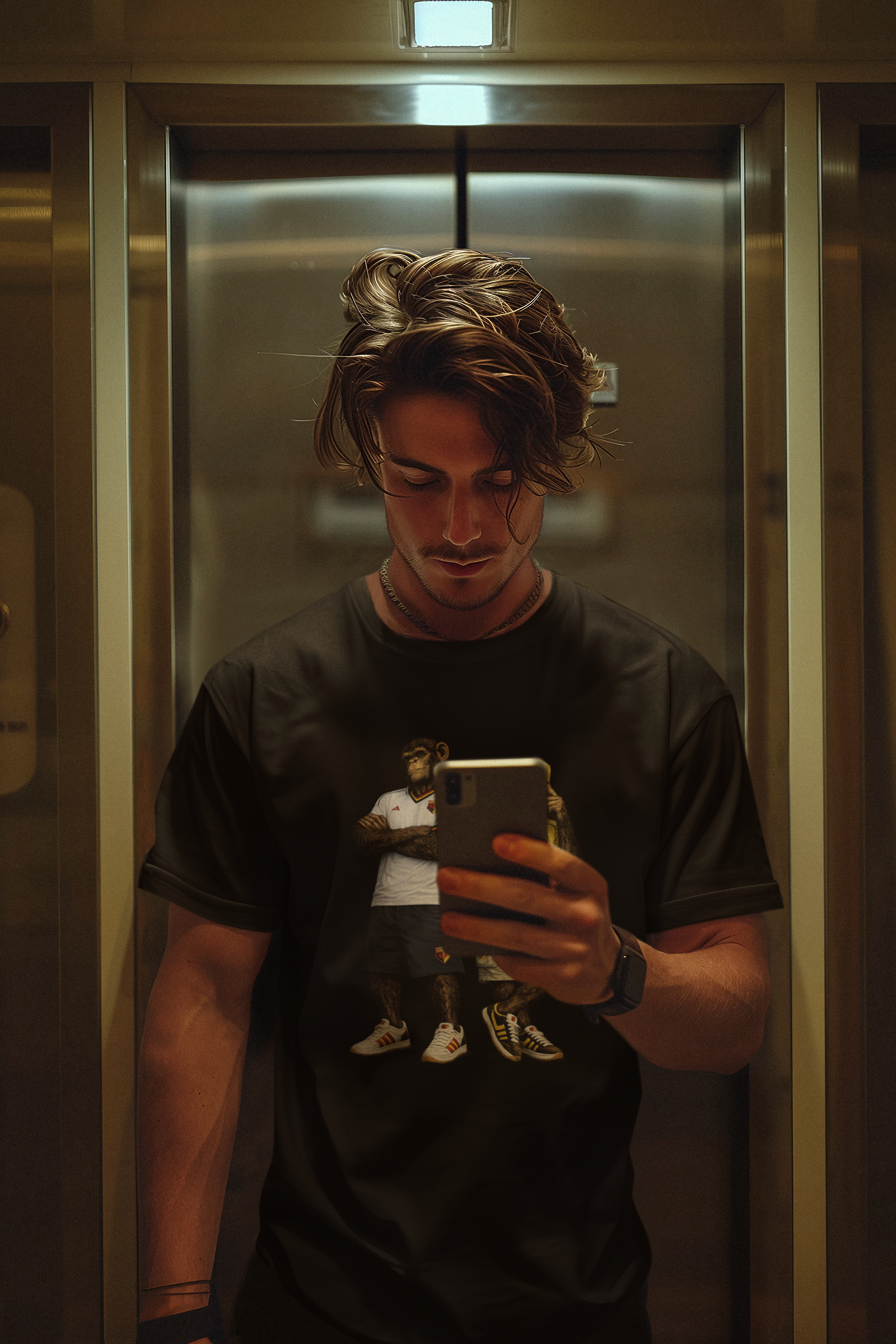 A man in a black Watford t-shirt taking a mirror selfie inside a metallic elevator lobby.