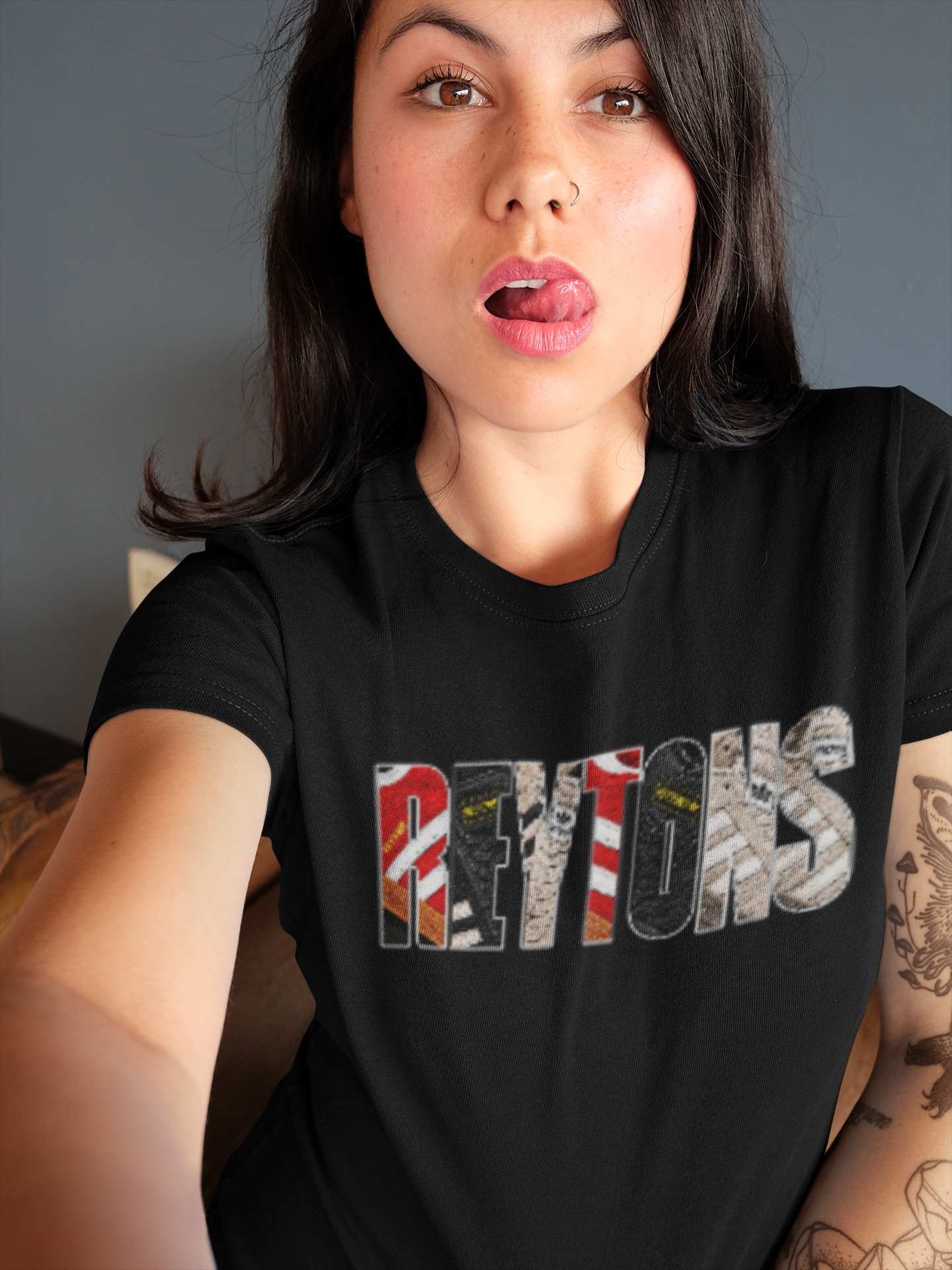A woman wearing a black Reytons t-shirt with a graphic spelling 'Reytons' filled with various flag patterns, photographed in a casual indoor setting.
