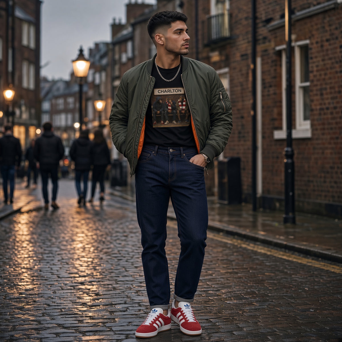 A man stands on a wet cobblestone street wearing an olive green bomber jacket over a black graphic CHARLTON t-shirt, dark blue jeans, and red low-top sneakers. The background shows blurred brick buildings and pedestrians.
